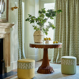 Living room with a round wooden table, potted plant, and two ottomans and oakleaf embroidery curtains on a green background.