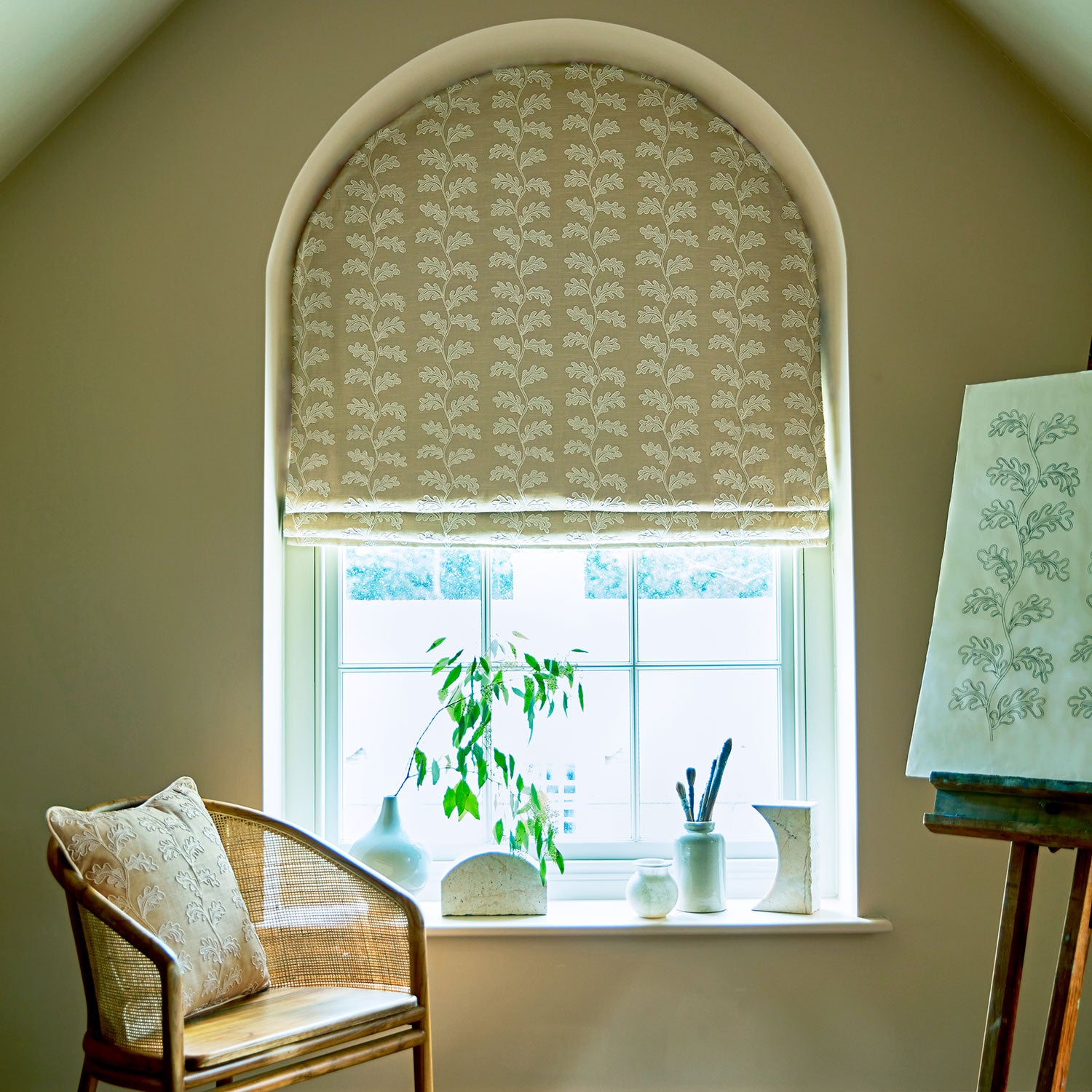 Decorative blind with taupe oakleaf embroidery pattern with a chair and plants.