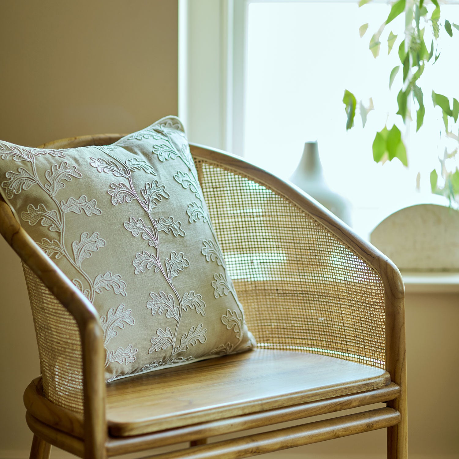 Wicker chair with a embroidery oakleaf pillow in a bright room with plants.