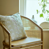 Wicker chair with a embroidery oakleaf pillow in a bright room with plants.