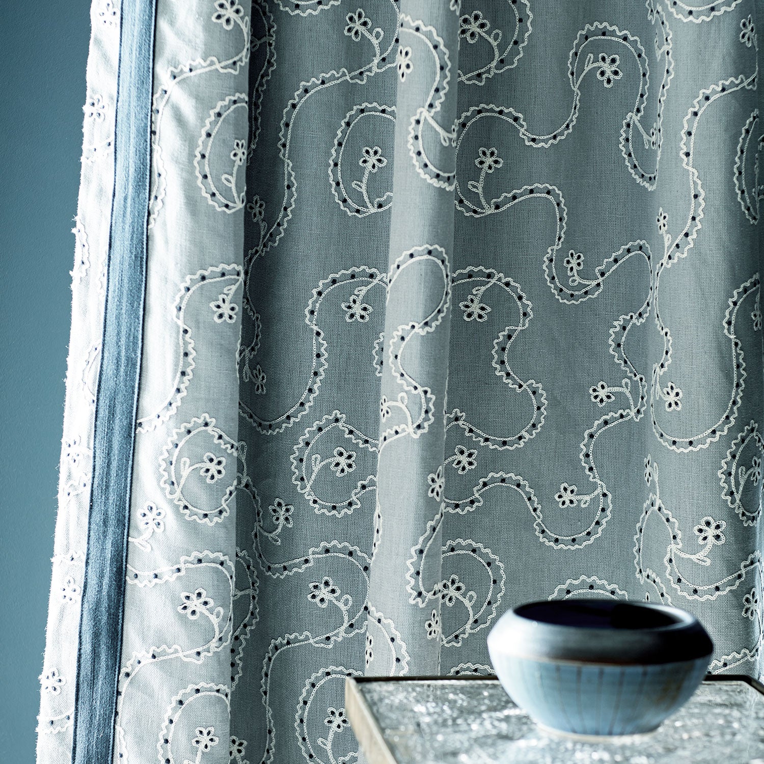 Embroidered blue curtain with a ceramic bowl on a table.