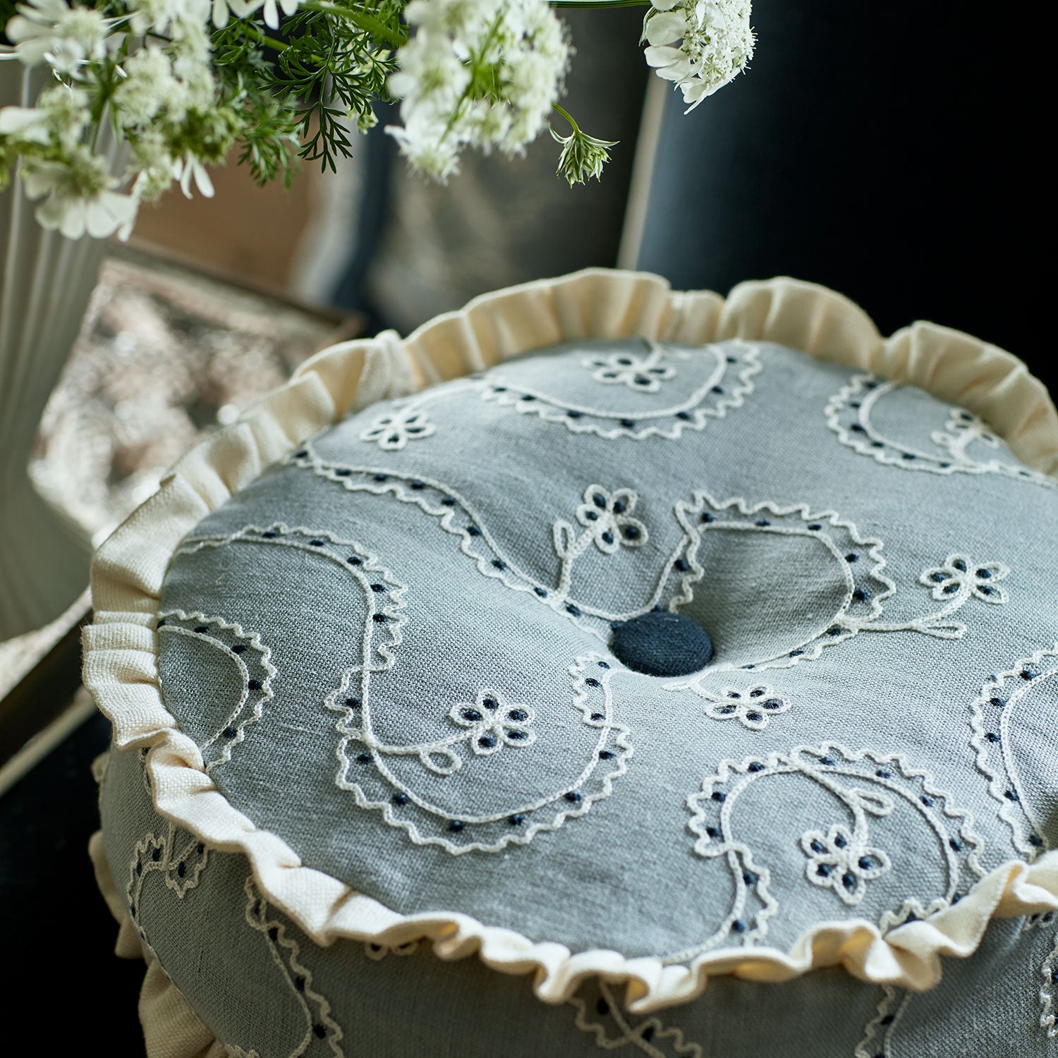 Decorative embroidered cushion with trim on a table, surrounded by white flowers.