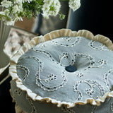 Decorative embroidered cushion with trim on a table, surrounded by white flowers.
