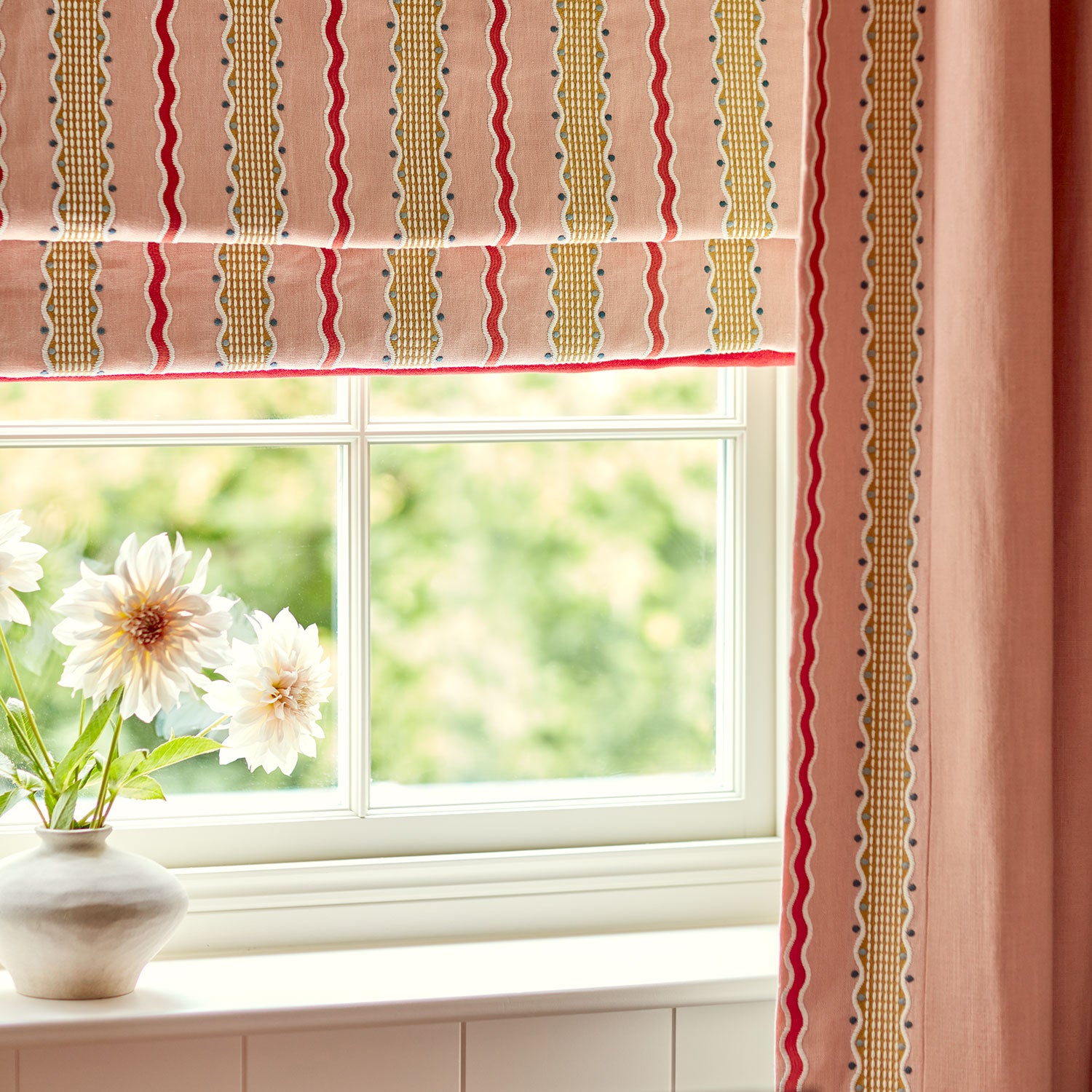Window with pink embroidered roman blind and curtains, featuring a vase with flowers on a windowsill.